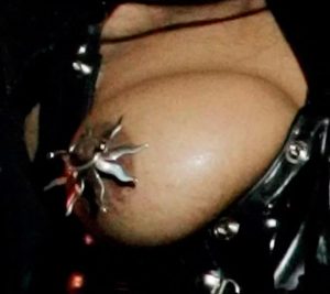 A close-up of a Janet's bare breast, covered only with a decorative, metallic sun-shaped nipple shield attached to her nipple with a piercing.