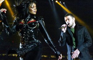 Janet Jackson in a black, shiny costume dances while Justin Timberlake in dark clothing sings into a microphone on stage under bright spotlights, with a large audience in the background.