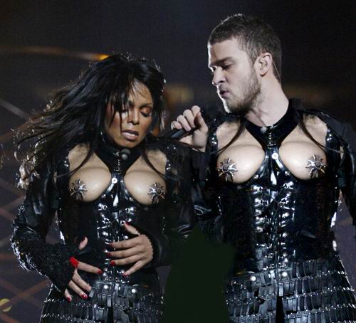 Janet Jackson and Justin Timberlake on stage wearing black leather outfits with metallic accents. Both of her breasts are exposed, both with the nipple shields. He is wearing the exact same outfit with the same exposed breasts, also with the same nipple shields, as he other sings into a microphone. The scene appears intense and dramatic.